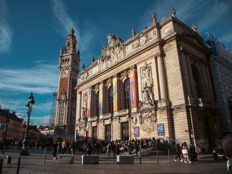 Grand’Place de Lille avec l’Opéra de Lille au premier plan, architecture néoclassique ornée de statues et de colonnes, et le Beffroi de la Chambre de Commerce en arrière-plan sous un ciel bleu éclatan