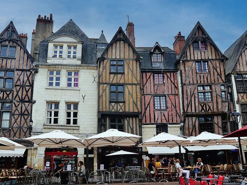 Façades de maisons à colombages colorées et typiques de l'architecture médiévale, alignées sur une place animée. Terrasse de café avec parasols blancs et clients attablés en plein air. Ciel bleu en ar