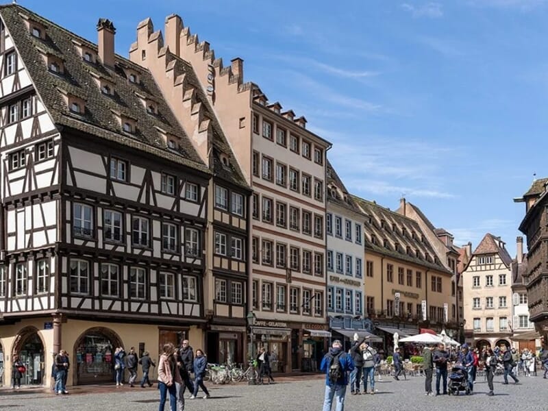 Place animée de Strasbourg avec maisons traditionnelles à colombages et bâtiments historiques, sous un ciel bleu ensoleillé