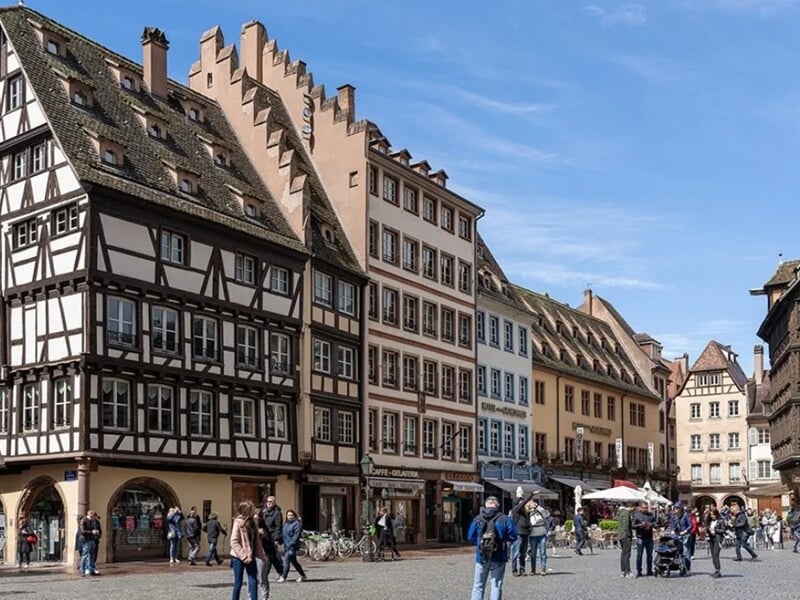 Place animée de Strasbourg avec maisons traditionnelles à colombages et bâtiments historiques, sous un ciel bleu ensoleillé