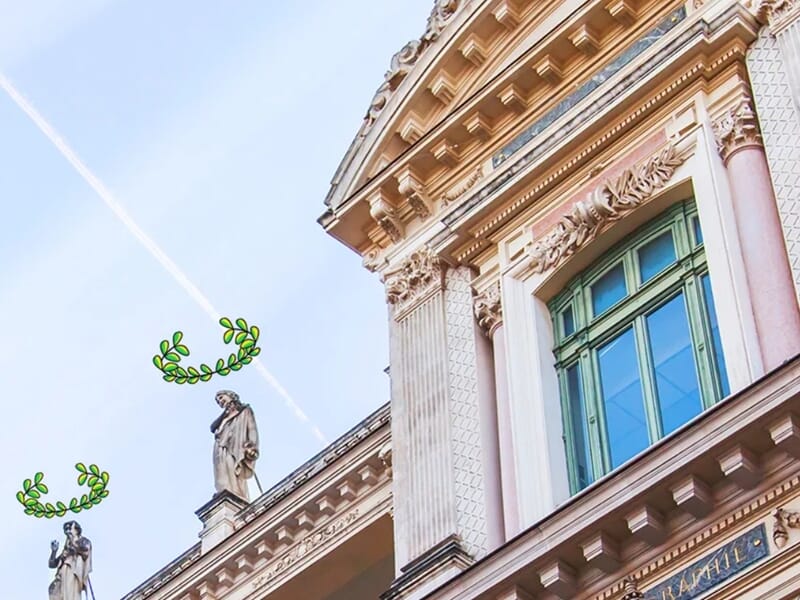 Façade néoclassique d’un bâtiment orné de statues et décorations sculptées, avec deux statues coiffées numériquement de couronnes de lauriers vertes