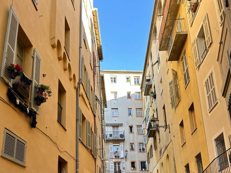 Ruelle étroite bordée d’immeubles aux façades pastel et volets typiques, sous un ciel bleu dégagé