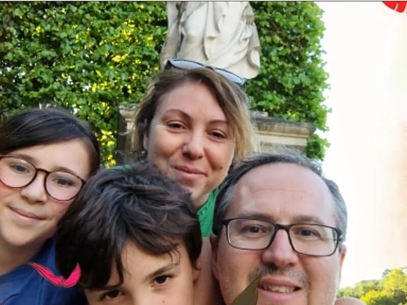 Photo d’une famille souriante composée de deux adultes et deux enfants, posant dans un jardin devant une statue, avec un décor de feuilles colorées en surimpression