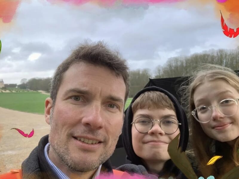 Selfie d’un père avec deux adolescents souriants portant des lunettes, dans un parc au ciel nuageux avec des éléments graphiques de feuilles colorées en bordure de photo
