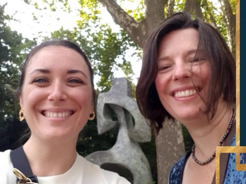 Selfie de deux femmes souriantes devant une sculpture moderne et des arbres dans un parc.