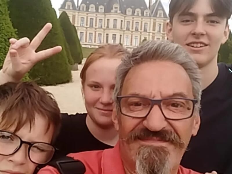 Selfie d’un homme et trois adolescents souriants devant un château avec des haies taillées en cône.