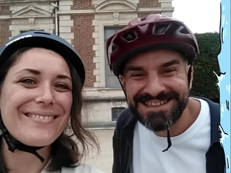 Selfie d’une femme et d’un homme souriants portant des casques de vélo, devant un bâtiment en briques.
