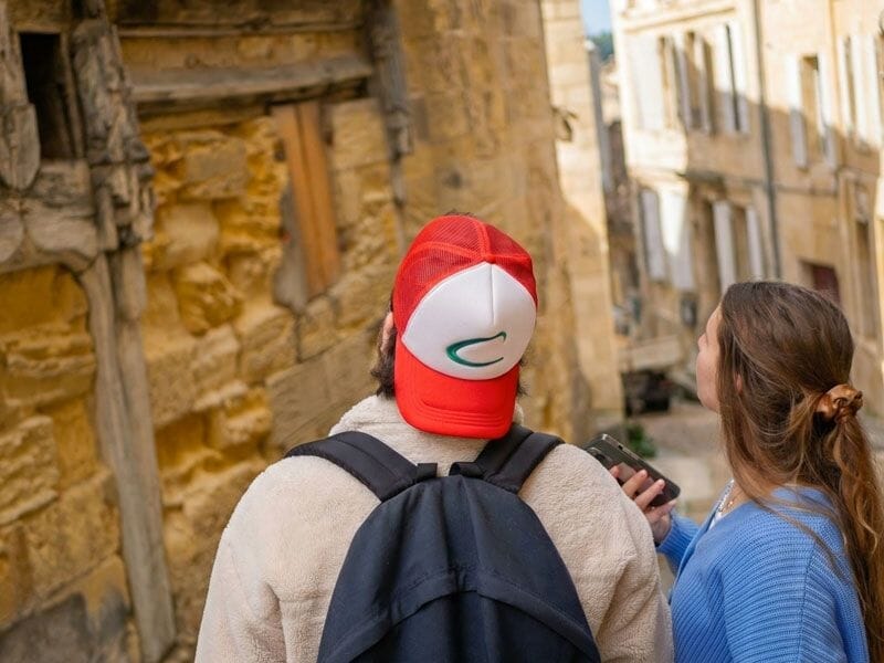 Deux jeunes regardent un téléphone en marchant dans une ruelle ancienne aux murs en pierre jaune.