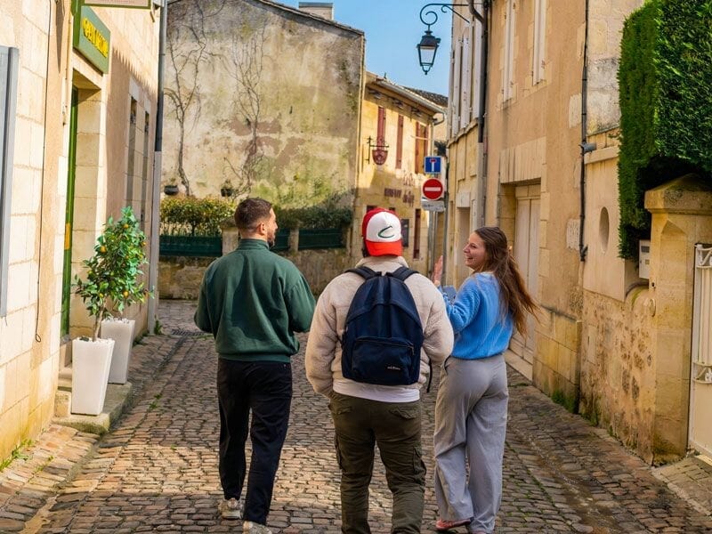 Trois personnes marchent dans une ruelle étroite de Saint-Émilion sous un ciel bleu clair.