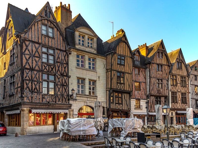 Façades anciennes à colombages dans une rue piétonne avec terrasses de café et chaises empilées.