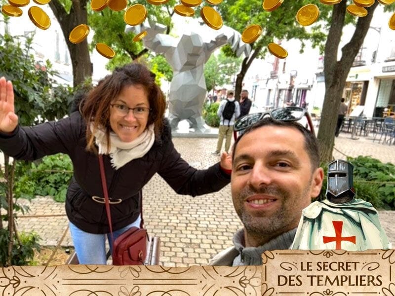 Deux personnes souriantes en extérieur avec une statue moderne et un panneau du jeu d'évasion 'Le secret des Templiers'.