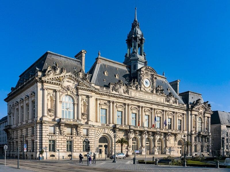 Bâtiment historique imposant avec horloge et sculptures, sous un ciel bleu clair sans nuages.