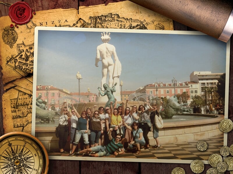 Groupe d’une vingtaine de personnes posant joyeusement devant la Fontaine du Soleil à Nice, dominée par la statue d’Apollon.