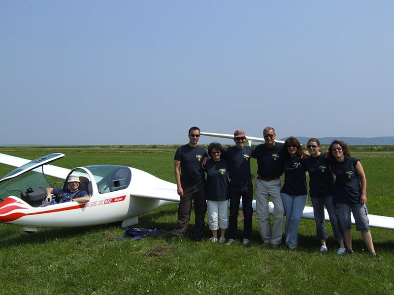 Un groupe de personnes posant à côté d'un planeur blanc sur une pelouse verte, avec un pilote assis dans le cockpit.