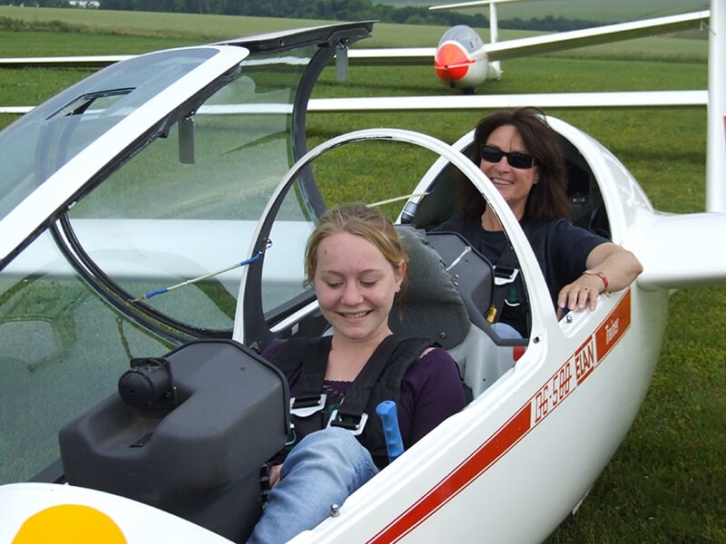 Deux femmes dans le cockpit d'un planeur, souriantes et prêtes à voler, sur une pelouse verte avec un autre planeur en arrière-plan.