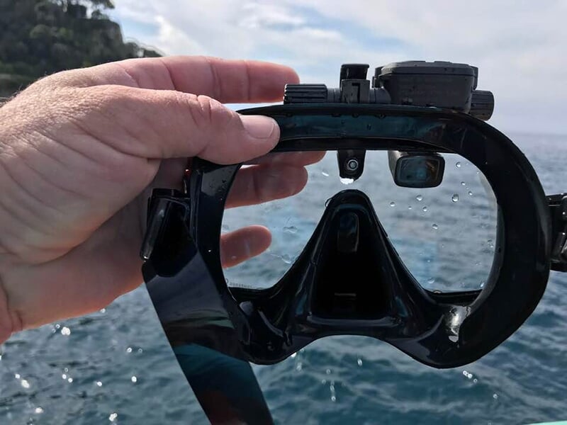 Une main tenant un masque de plongée noir, avec la mer et un paysage côtier flou en arrière-plan.