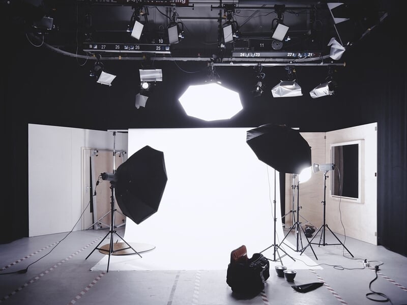 Studio de photographie équipé de multiples sources lumineuses, dont des softbox et des projecteurs suspendus, dirigés vers un fond blanc. Deux grands parapluies noirs sont installés sur des trépieds p