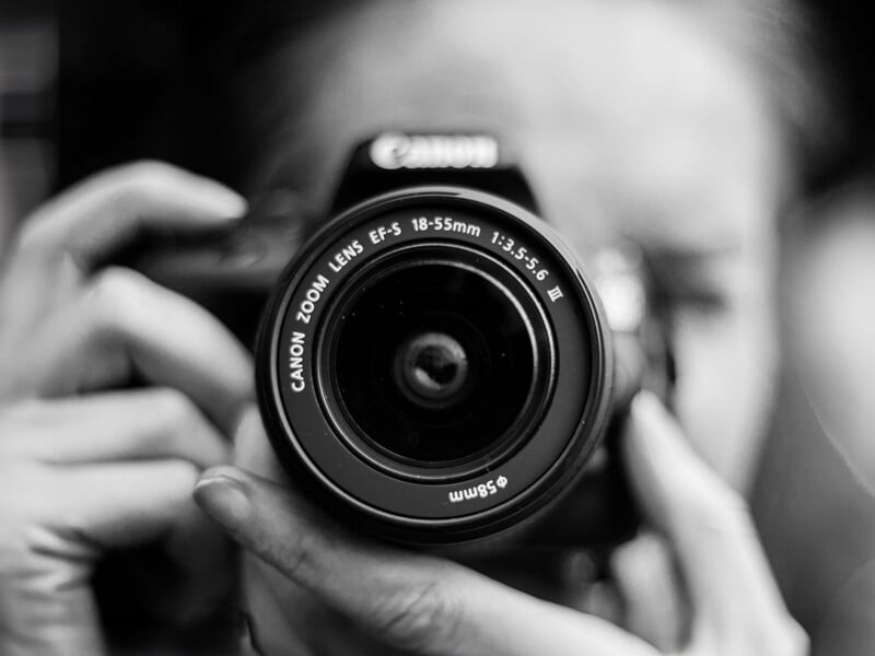 Gros plan sur un photographe tenant un appareil photo reflex Canon avec un objectif 18-55mm. L’image en noir et blanc met en valeur les détails de l’objectif et les mains du photographe, créant une pe