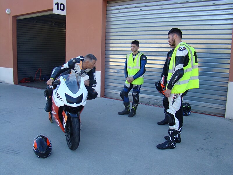 Motard assis sur une moto de course blanche discutant avec deux autres personnes portant des combinaisons et des gilets fluorescents lors d'un stage de moto sur circuit 