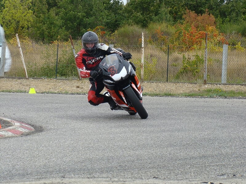Stage moto avec motard en combinaison rouge prenant un virage incliné sur une moto de course sur un circuit.