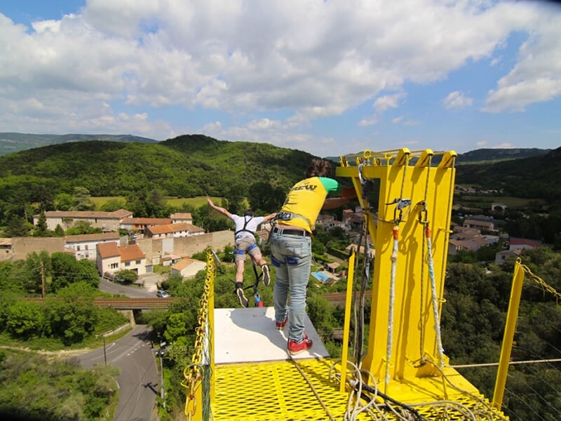  Personne effectuant un saut à l'élastique depuis une plateforme installée sur un viaduc, avec un moniteur en arrière-plan et un paysage verdoyant.