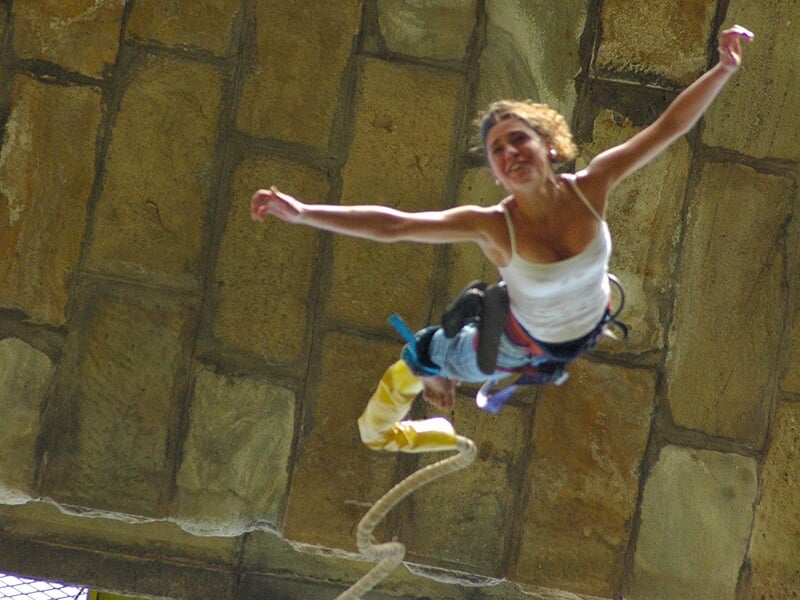 Femme en plein saut à l'élastique sous un viaduc, les bras ouverts et un sourire exprimant l'adrénaline.