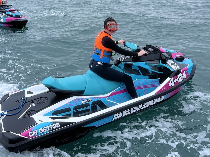 Une personne seule sur une motomarine colorée bleu, rose et blanc, portant un gilet de sauvetage orange et des lunettes de protection, en mer.