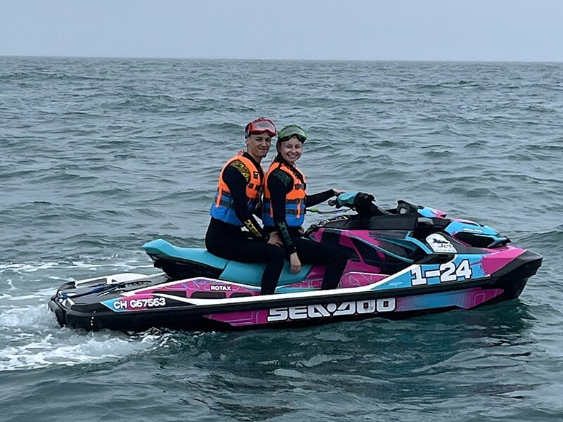 Deux personnes assises sur une motomarine rose, bleu et blanc, souriantes et portant des gilets de sauvetage orange.