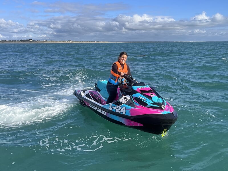 Une femme souriante seule sur une motomarine rose et bleu, naviguant sur une mer calme avec un paysage côtier au loin.