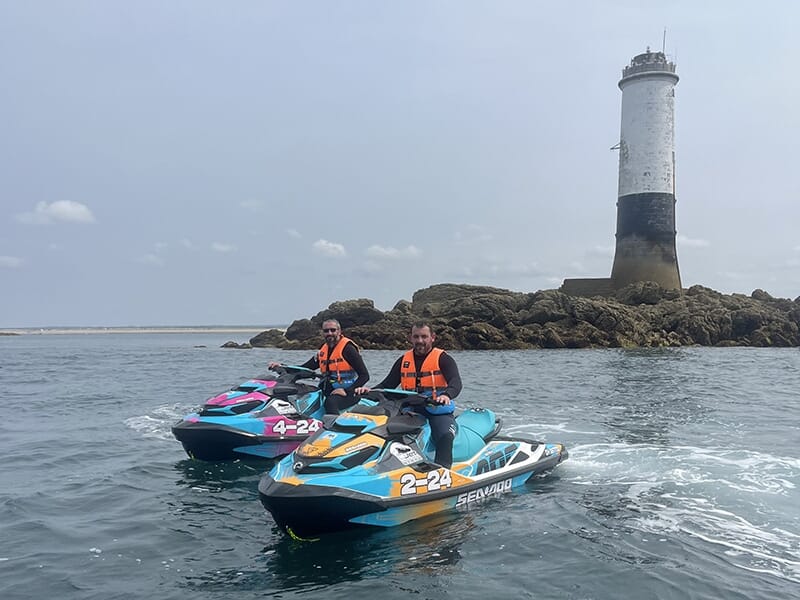 Deux motomarines près d’un phare en mer, avec des rochers visibles en arrière-plan, les pilotes portant des gilets orange.