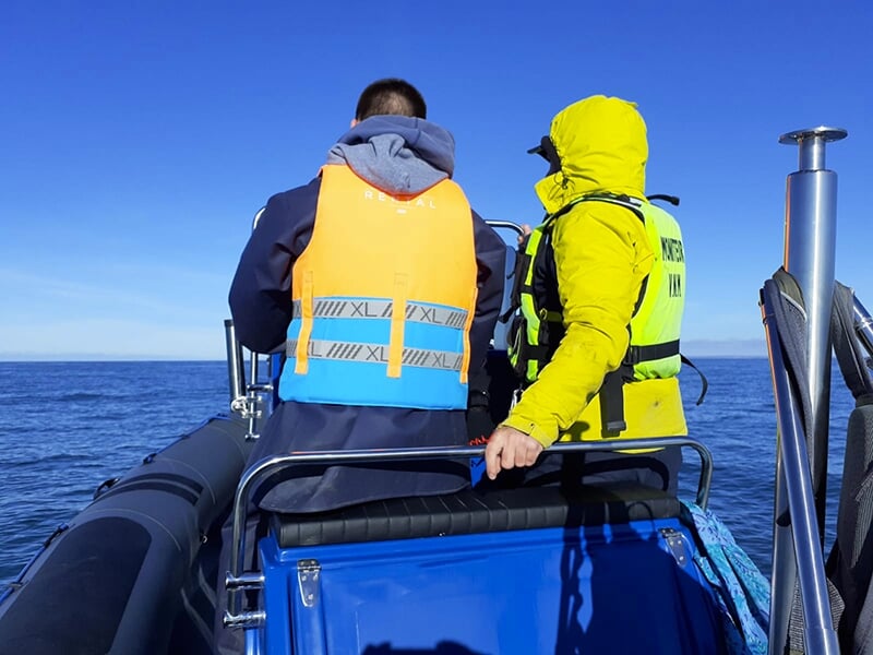 Deux personnes en gilets de sauvetage, dont l'une vêtue d'un anorak jaune, à bord d'un bateau pneumatique naviguant en mer sous un ciel dégagé.