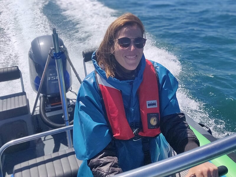 Une femme souriante portant une veste bleue et un gilet de sauvetage rouge, assise à bord d'un bateau à moteur rapide en pleine navigation sur une mer calme