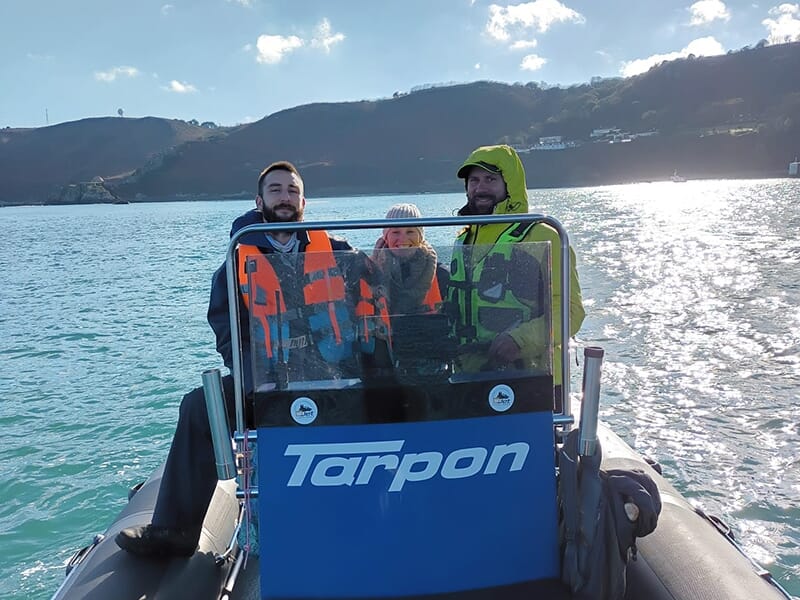 Trois personnes portant des gilets de sauvetage, à bord d'un bateau pneumatique avec le logo 