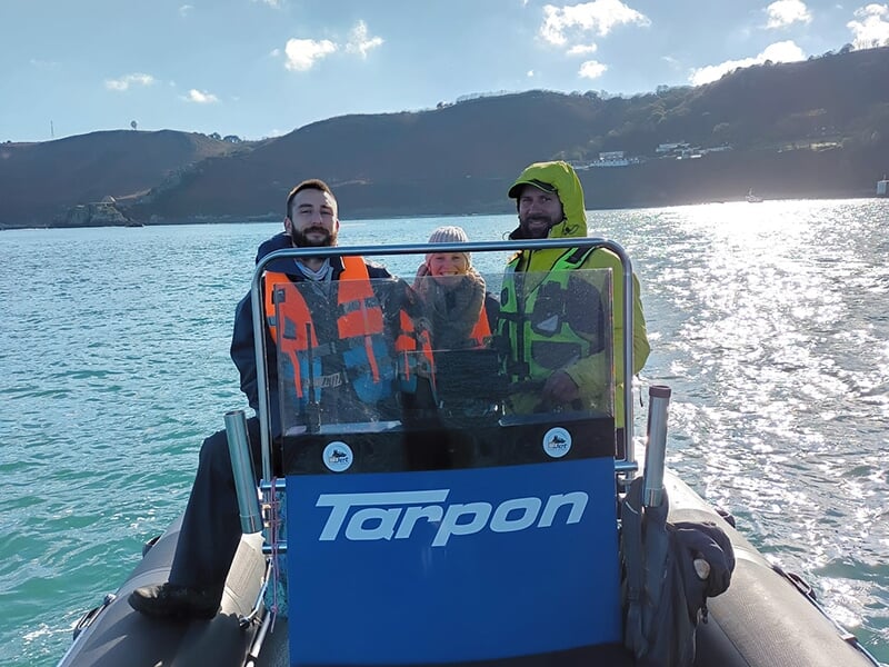 Trois personnes portant des gilets de sauvetage, à bord d'un bateau pneumatique avec le logo 