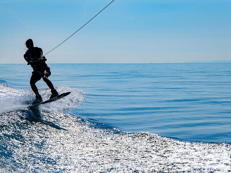 Un pratiquant de wakeboard glissant sur l'eau, tiré par une corde, avec un ciel bleu dégagé et des reflets scintillants sur la mer.