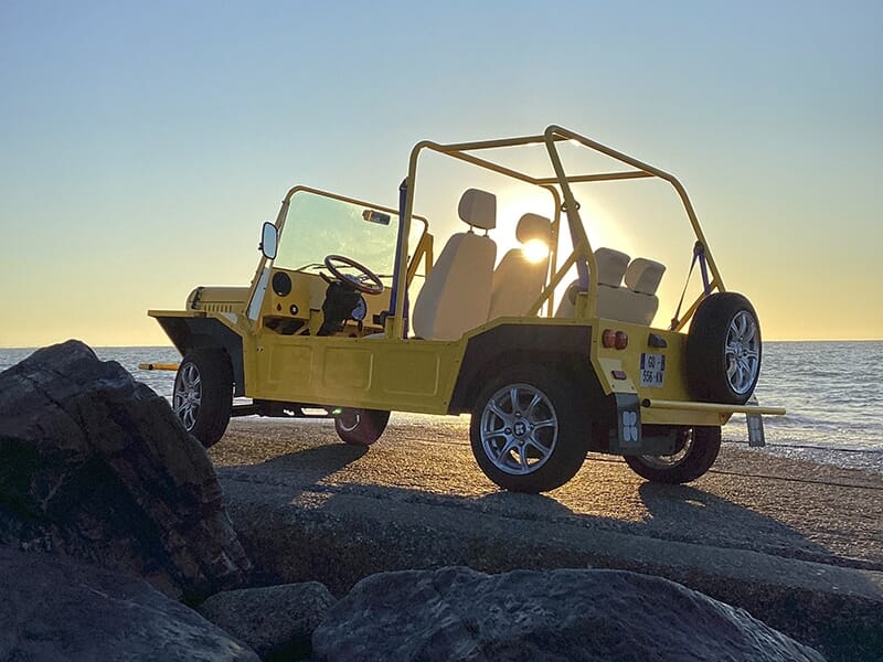 Voiture jaune décapotable avec le soleil couchant visible à travers sa structure, garée près de la mer.