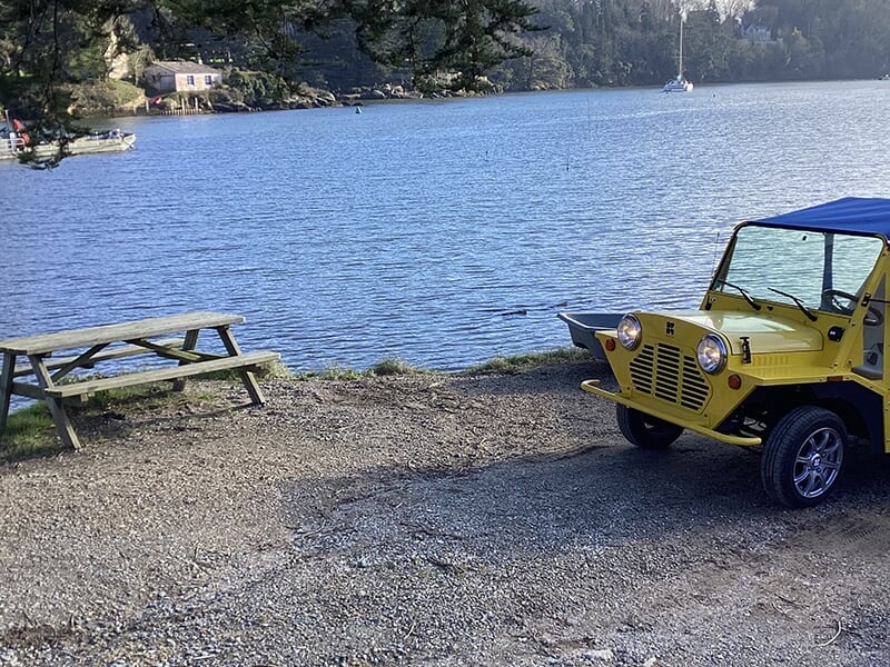 Voiture jaune décapotable garée près d’un espace pique-nique au bord de l’eau.