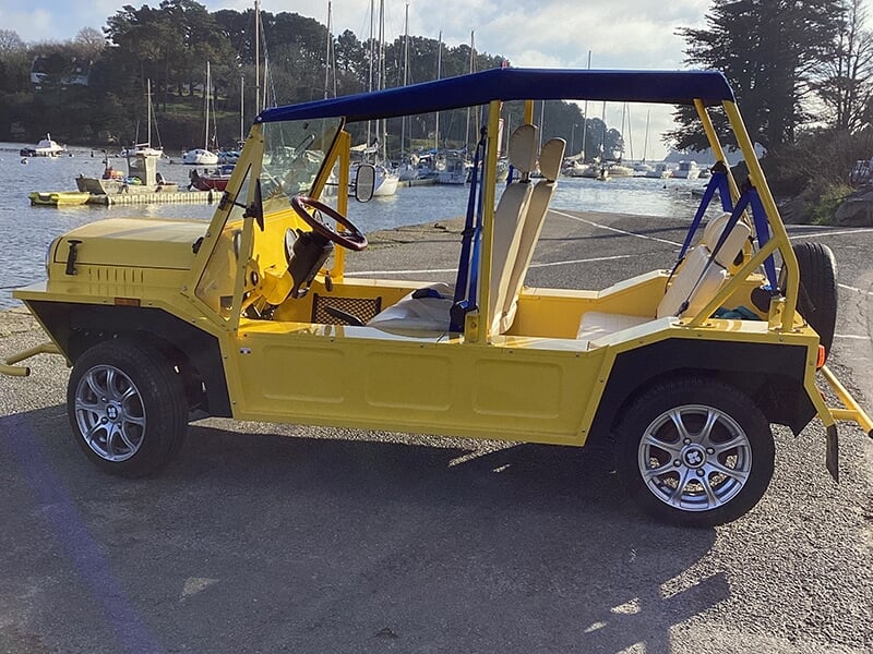 Voiture décapotable jaune vintage garée près d'un port avec des voiliers et arbres en arrière-plan sous un ciel clair.