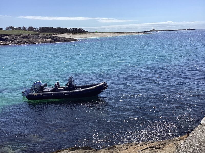 Un bateau pneumatique noir flottant sur une eau turquoise et scintillante, avec une plage et un paysage côtier en arrière-plan.