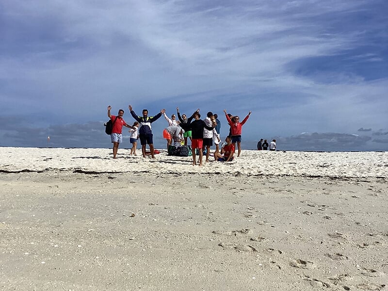 Un groupe de personnes debout sur une plage de sable blanc, levant les bras dans un geste joyeux.
