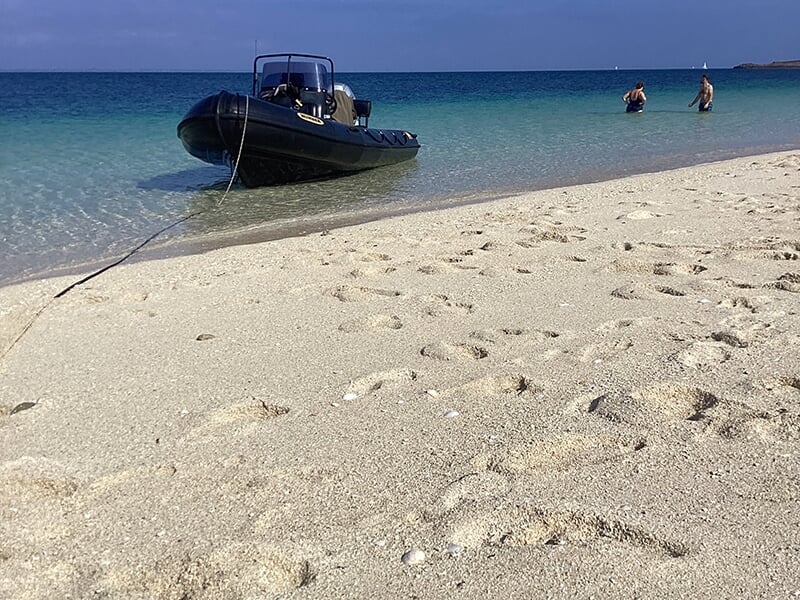 Une plage de sable fin avec des empreintes, un bateau noir au bord de l'eau turquoise et deux personnes se baignant.
