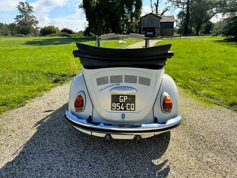 Arrière d'une Volkswagen Coccinelle décapotable blanche avec capote repliée, vue sur une allée gravillonnée.