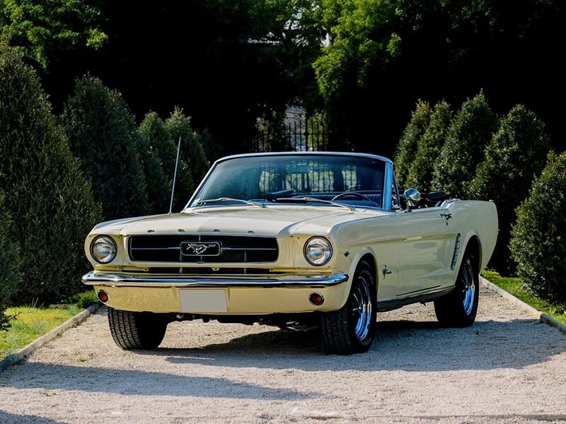 Vue avant d'une Ford Mustang décapotable crème, garée sur une allée gravillonnée entourée de haies.