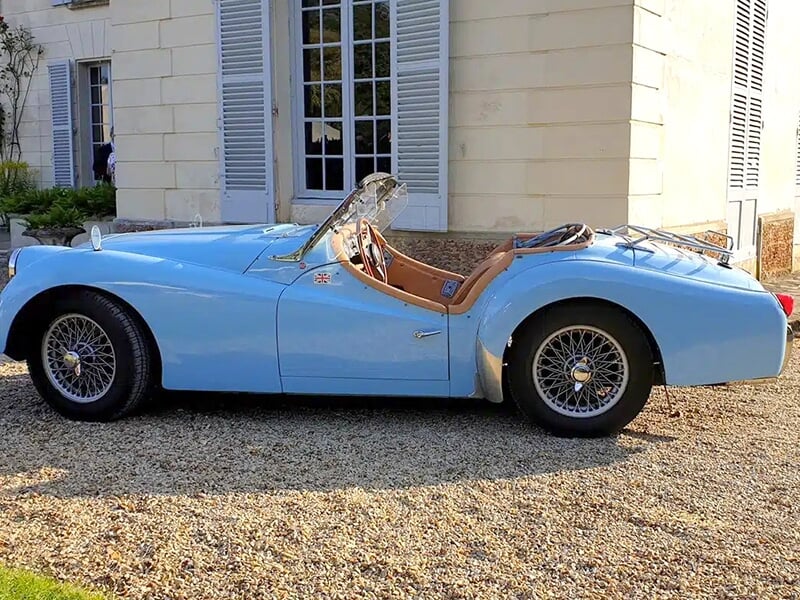 Profil d'une Triumph TR3 bleue décapotable garée devant une maison avec des volets blancs.