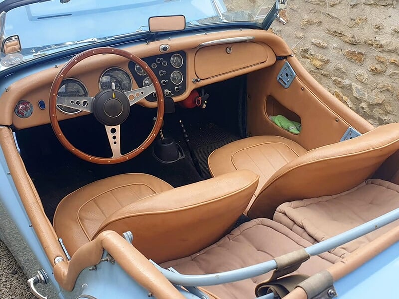 Intérieur d'une Triumph TR3 bleue avec un volant en bois et des sièges en cuir beige.