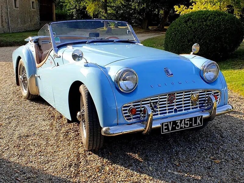 Vue avant d'une Triumph TR3 bleue décapotable avec des phares ronds et une grille chromée, garée sur du gravier.