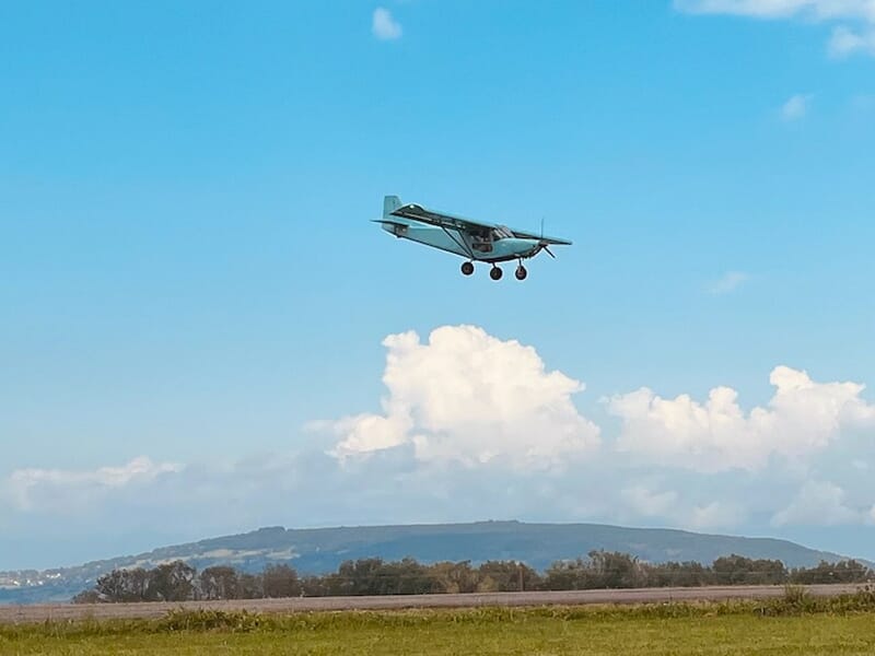 Un avion léger en plein vol sous un ciel bleu clair avec des nuages blancs et un paysage vallonné en arrière-plan.