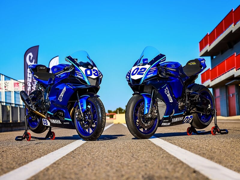 Deux motos Yamaha bleues n°03 et n°02 stationnées côte à côte sur une piste, prêtes pour une course.