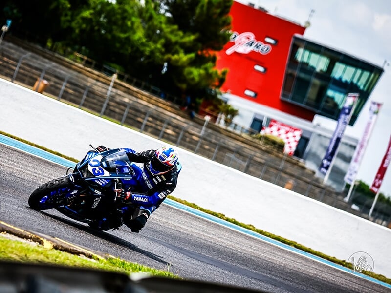 Motard sur une Yamaha n°03 dans un virage, avec un bâtiment et des tribunes visibles en arrière-plan.