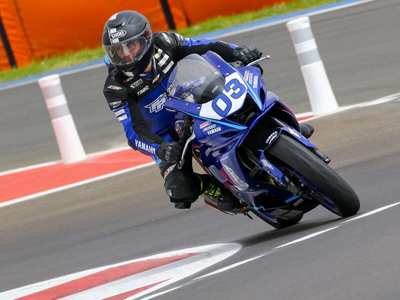 Motard sur une Yamaha n°03 en train de franchir un virage avec un fond de piste et des éléments de sécurité visibles.